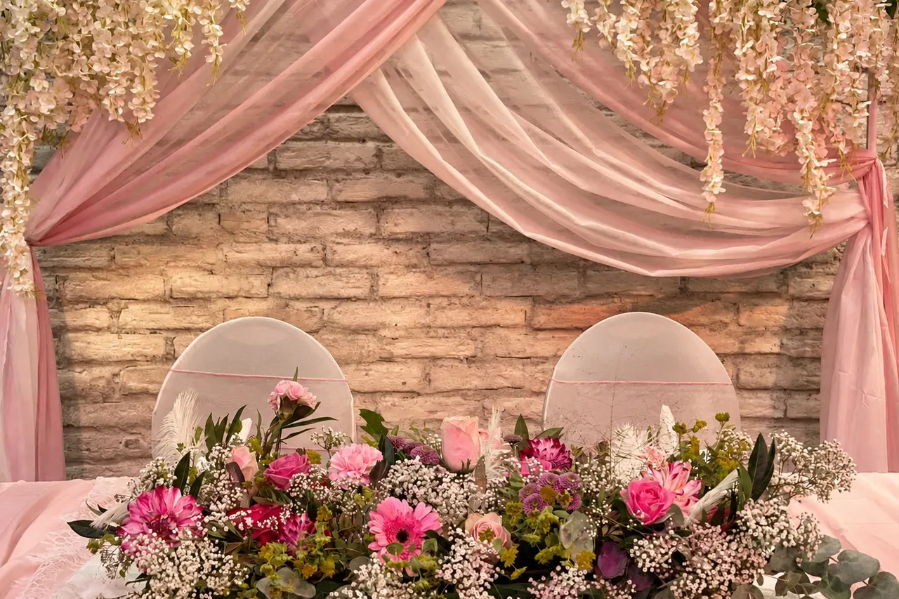 Brauttisch vor einer Backsteinwand mit rosa Stoffdraperie, Blumengirlanden und einem Blumenarrangement